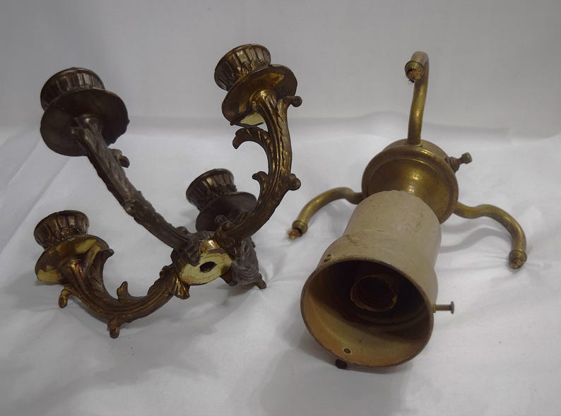 May include: Two antique brass candle holders, one with three arms and the other with a single arm. Both have ornate designs and are in good condition.