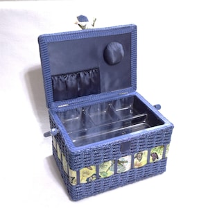 May include: A blue woven sewing basket with a hinged lid. The interior features a dark blue fabric lining with a pocket and a circular pin cushion. A clear plastic tray with multiple compartments sits inside. The exterior has a butterfly print band.