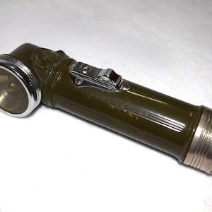May include: An olive-drab vintage flashlight with a silver-colored bezel and end cap. The flashlight has a right-angle head and a switch on the top. The body has a ribbed design.