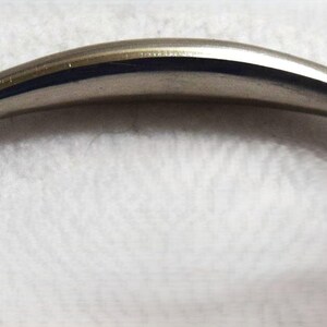 May include: A metallic silver-toned cabinet handle with a smooth, arched design. The handle features a curved center section that tapers toward two flat, wide mounting bases on each end, each containing a single circular screw hole for installation.