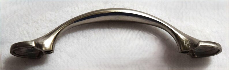 May include: A metallic silver-toned cabinet handle with a smooth, arched design. The handle features a curved center section that tapers toward two flat, wide mounting bases on each end, each containing a single circular screw hole for installation.