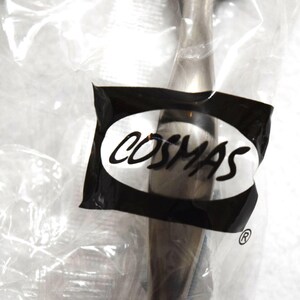 May include: A metallic cabinet pull with a brushed finish, encased in a clear plastic bag. A black label on the front of the packaging features the white text COSMAS inside an oval shape, with a registered trademark symbol located below the label.