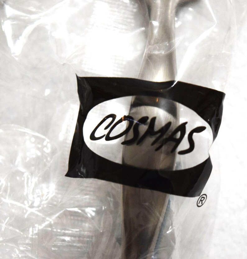 May include: A metallic cabinet pull with a brushed finish, encased in a clear plastic bag. A black label on the front of the packaging features the white text COSMAS inside an oval shape, with a registered trademark symbol located below the label.