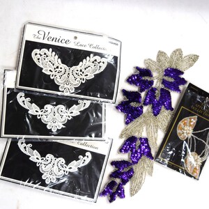 May include: Several lace appliques and embellishments are displayed on a white surface. Three packages of white lace necklines from "The Venice Lace Collection" are visible. A silver and purple sequined floral applique and a package of beaded appliques are also present.