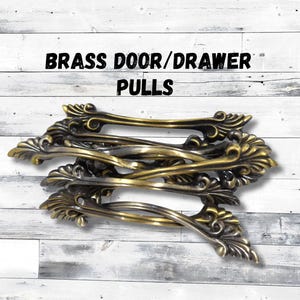 May include: A collection of brass door and drawer pulls. The pulls have an ornate, vintage design with a curved handle and decorative floral accents. The text "BRASS DOOR/DRAWER PULLS" is displayed above the pulls.