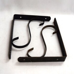 May include: Two black metal shelf brackets with an S-shaped design. The brackets have a rectangular base with pre-drilled holes for mounting. The curved support arms extend from the base, ending in a small hook. The brackets are made of a dark, possibly aged, metal.