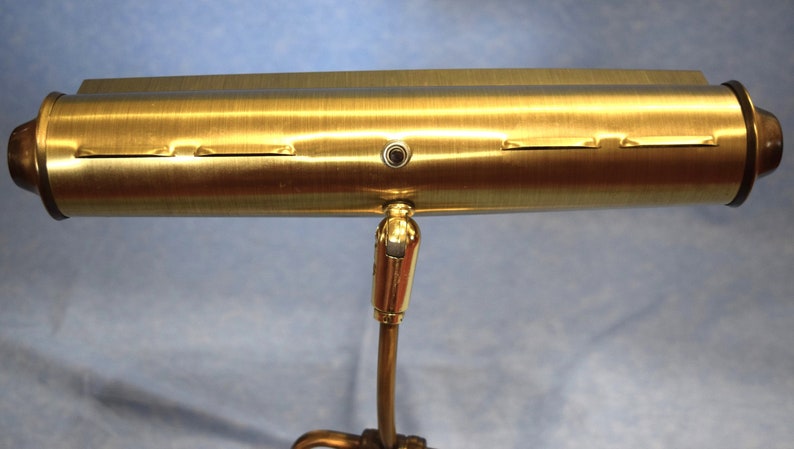 K&ouml;nnte beinhalten: Eine Schreibtischlampe aus Messing mit einem zylindrischen Schirm und einem flexiblen Arm. Der Schirm hat eine geb&uuml;rstete Oberfl&auml;che und drei Schlitze f&uuml;r Gl&uuml;hbirnen.