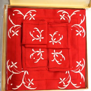 May include: A red linen table set with white floral embroidery. The set includes a large square tablecloth and four matching square napkins, all neatly arranged in a box. The white embroidery creates a striking contrast against the red fabric.