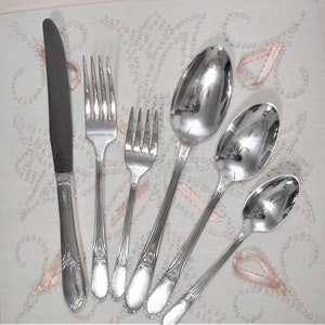 May include: A set of silver flatware, including a knife, fork, and spoons. The flatware is arranged on a white background with pink accents.