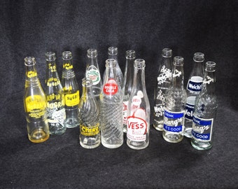 May include: A collection of vintage glass soda bottles with various labels, including "Nugra", "Chero", "Double Cola", "Vess", "Welch's", and "Barg's".