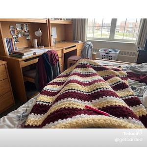 May include: A handmade crochet blanket with alternating stripes of burgundy, cream, and gold. The blanket is draped over a bed in a dorm room setting. A pink crochet hook rests on the blanket. The room includes a desk, chair, and window.