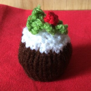 May include: A knitted Christmas pudding ornament with a white icing top and a green holly sprig with a red berry.