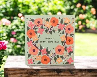 May include: A square Mother's Day card with a light green background. The card features a floral heart design in shades of orange, pink, and purple, with the words "HAPPY MOTHER'S DAY" in the center. The card is set on a wooden surface.