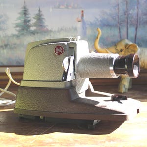 May include: Vintage silver and brown slide projector with a black lens. The projector has a textured finish and a power cord. A small red and white logo is visible on the side. An elephant figurine and a framed painting are in the background.