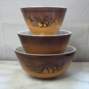 Puede incluir: Un conjunto de tres cuencos de mezcla vintage apilados. Los cuencos son marrones con un degradado naranja. Cada cuenco presenta una ilustración negra de uvas y otras frutas. Los interiores son blancos. Un clásico juego de utensilios de cocina.
