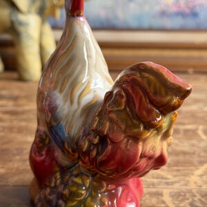 May include: A ceramic rooster figurine featuring a glossy glaze with a color palette of cream, deep red, brown, and hints of blue. The sculpture depicts the bird in a standing position with a prominent red comb and textured plumage details.