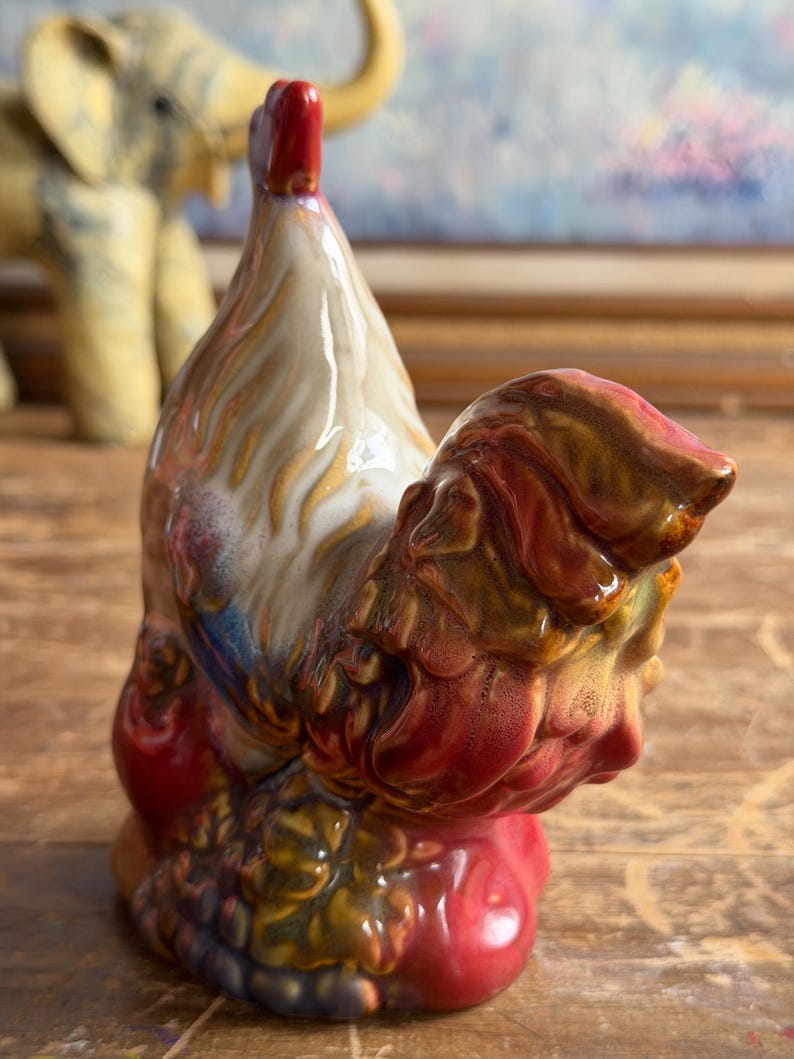 May include: A ceramic rooster figurine featuring a glossy glaze with a color palette of cream, deep red, brown, and hints of blue. The sculpture depicts the bird in a standing position with a prominent red comb and textured plumage details.