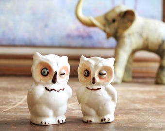 May include: Two ceramic owl figurines with winking eyes and brown beaks. The owls are white with brown and peach-colored accents. An elephant figurine is in the background. The figurines are on a wooden surface.