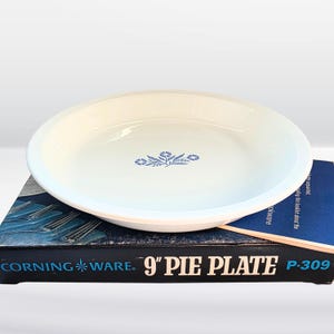 May include: A white Corning Ware pie plate with a blue floral design in the center. The pie plate is 9 inches in diameter and sits on top of its original box. The box is black with white text that reads "CORNING WARE. 9" PIE PLATE P-309."