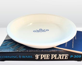 May include: A white Corning Ware pie plate with a blue floral design in the center. The pie plate is 9 inches in diameter and sits on top of its original box. The box is black with white text that reads "CORNING WARE. 9" PIE PLATE P-309."