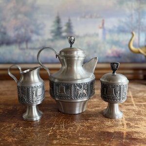 May include: A three-piece pewter tea set consisting of a lidded teapot, a creamer, and a lidded sugar bowl. Each piece features embossed panels depicting Viking ships and figures, with small shield-shaped finials on the lids of the teapot and bowl.
