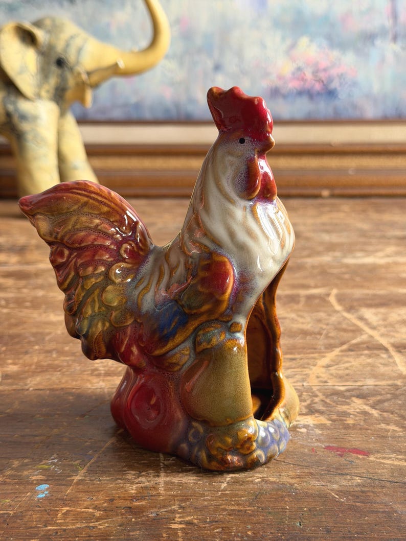 May include: A ceramic rooster figurine featuring a glossy, multi-colored glaze in shades of red, brown, cream, and blue. The rooster is depicted in a standing position with a prominent red comb, detailed feather textures, and a hollow base.