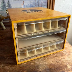 May include: A wooden storage cabinet with three clear plastic drawers, each featuring internal dividers. The top surface displays a circular DMC logo. The unit has a light brown wood grain finish and clear drawer fronts with the DMC brand name embossed.