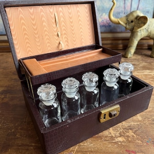 May include: A dark brown, textured rectangular box with a hinged lid and a gold-tone metal latch. The interior features a light wood-grain lining and a tiered tray holding five clear glass bottles with matching faceted glass stoppers.