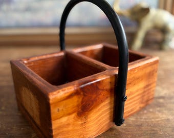 May include: A rectangular wooden caddy featuring two open compartments and a smooth, polished finish with visible wood grain. The caddy includes a single, arched black metal handle attached to the sides with small screws, providing a sturdy grip.