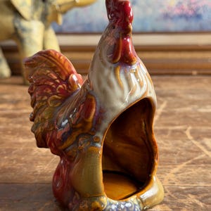 May include: A ceramic rooster figurine with a glossy, multi-colored glaze featuring shades of red, brown, tan, and blue. The rooster is depicted in a standing position with a hollowed-out front section, showcasing a smooth, reflective surface finish.
