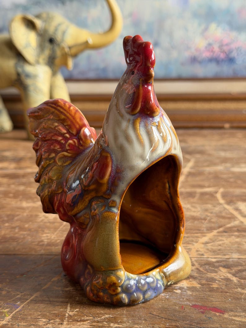 May include: A ceramic rooster figurine with a glossy, multi-colored glaze featuring shades of red, brown, tan, and blue. The rooster is depicted in a standing position with a hollowed-out front section, showcasing a smooth, reflective surface finish.
