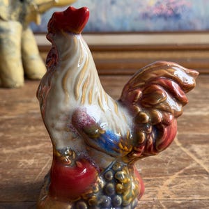 May include: A ceramic rooster figurine featuring a glossy glaze in shades of cream, red, brown, and blue. The rooster stands atop a base decorated with sculpted grapes and a red apple, with its head held high and a prominent red comb on top.