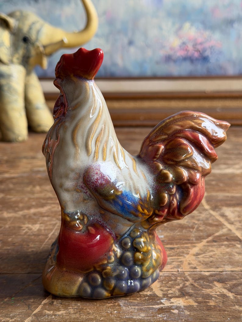 May include: A ceramic rooster figurine featuring a glossy glaze in shades of cream, red, brown, and blue. The rooster stands atop a base decorated with sculpted grapes and a red apple, with its head held high and a prominent red comb on top.