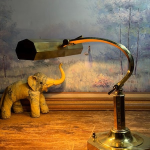 May include: A vintage brass desk lamp with an adjustable arm and a hexagonal shade. The lamp is positioned on a wooden surface, with a decorative elephant figurine nearby. The background features a framed landscape painting.