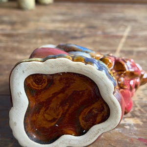May include: A ceramic figurine base with a glossy, marbled brown and amber glaze. The underside features a cream-colored, scalloped edge border surrounding the central glazed area, showcasing a smooth, reflective finish with visible swirling patterns.