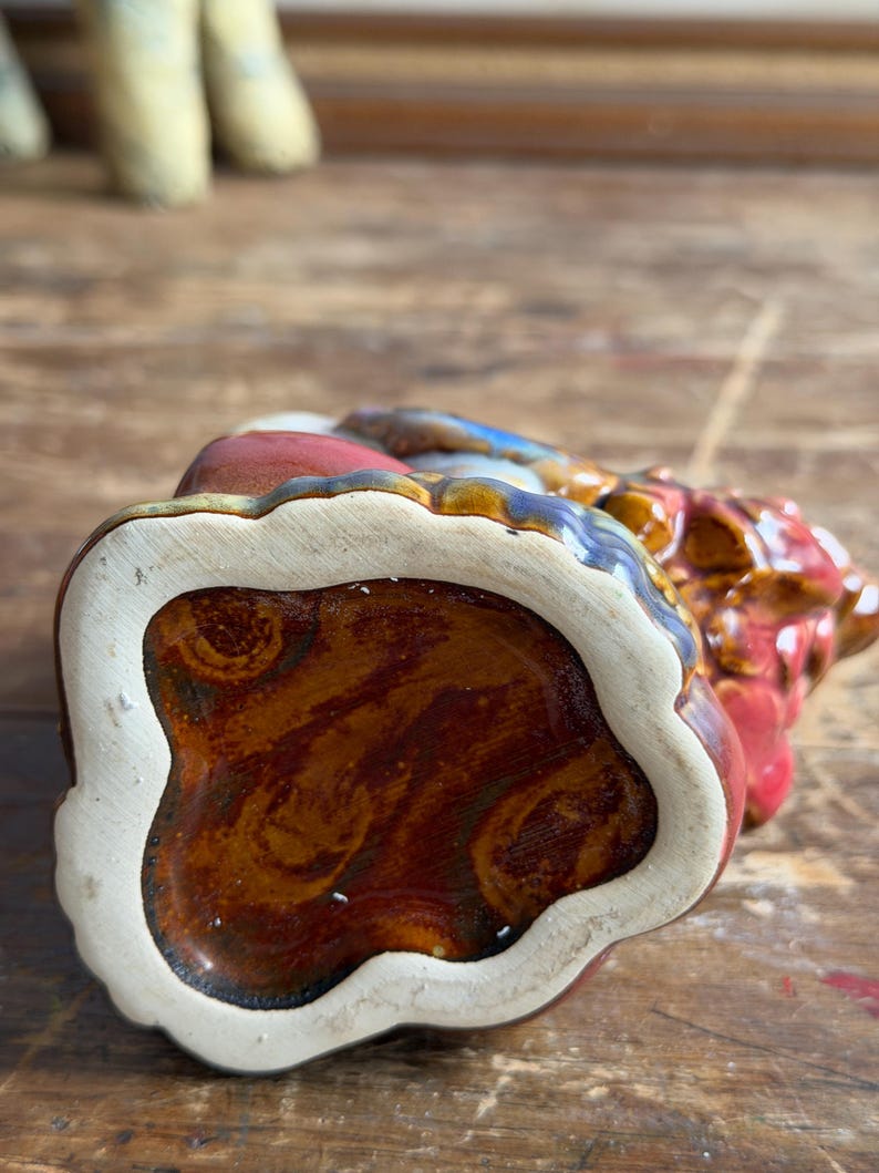 May include: A ceramic figurine base with a glossy, marbled brown and amber glaze. The underside features a cream-colored, scalloped edge border surrounding the central glazed area, showcasing a smooth, reflective finish with visible swirling patterns.