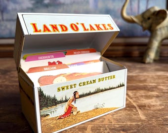 May include: A rectangular metal recipe box featuring the Land O'Lakes Sweet Cream Butter branding. The lid is open, revealing colorful tabbed dividers labeled Desserts, Main Dishes, and Breads. The box displays a vintage illustration of a person.