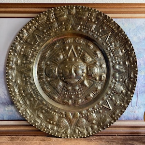 May include: A large, circular brass wall plaque featuring an embossed central face surrounded by concentric rings of intricate geometric patterns, stylized animal figures, and symbolic motifs, all rendered in a polished, metallic gold-toned finish.