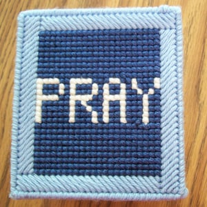 May include: A blue and white crocheted square with the word "PRAY" in white yarn.