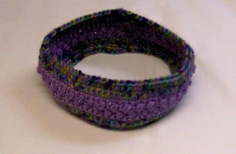 May include: A purple, green, and blue crocheted headband. The headband is made with a textured stitch and has a wide, comfortable fit.