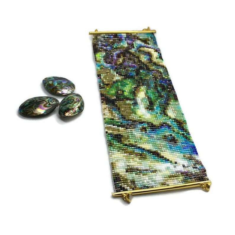 K&ouml;nnte beinhalten: Ein Armband aus Perlen mit einem farbenfrohen abstrakten Design in Gr&uuml;n-, Blau- und Braunt&ouml;nen. Das Armband besteht aus kleinen, quadratischen Perlen und hat einen goldenen Metallverschluss.