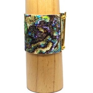 K&ouml;nnte beinhalten: Ein Armband aus Perlen mit einem farbenfrohen abstrakten Muster und einem goldenen Metallverschluss. Das Armband wird auf einem Holzst&auml;nder pr&auml;sentiert.