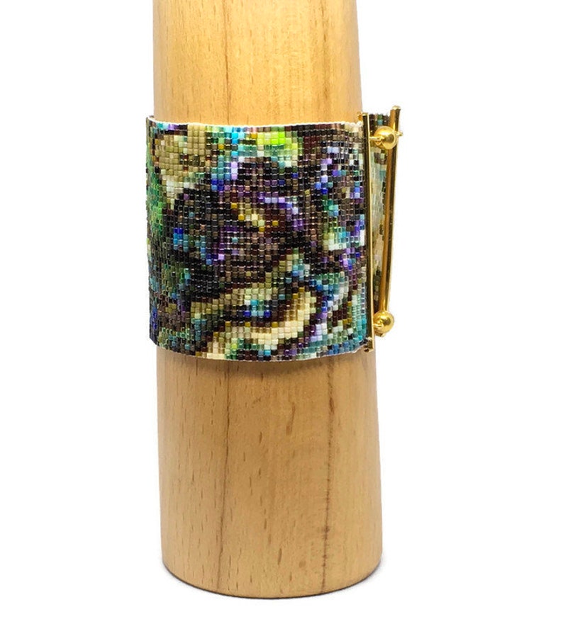 K&ouml;nnte beinhalten: Ein Armband aus Perlen mit einem farbenfrohen abstrakten Muster und einem goldenen Metallverschluss. Das Armband wird auf einem Holzst&auml;nder pr&auml;sentiert.
