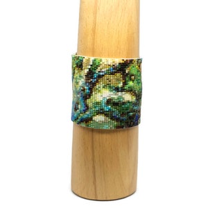 K&ouml;nnte beinhalten: Ein Armband aus Perlen mit einem farbenfrohen abstrakten Muster in Gr&uuml;n-, Blau- und Wei&szlig;t&ouml;nen. Das Armband wird auf einem Holzst&auml;nder pr&auml;sentiert.