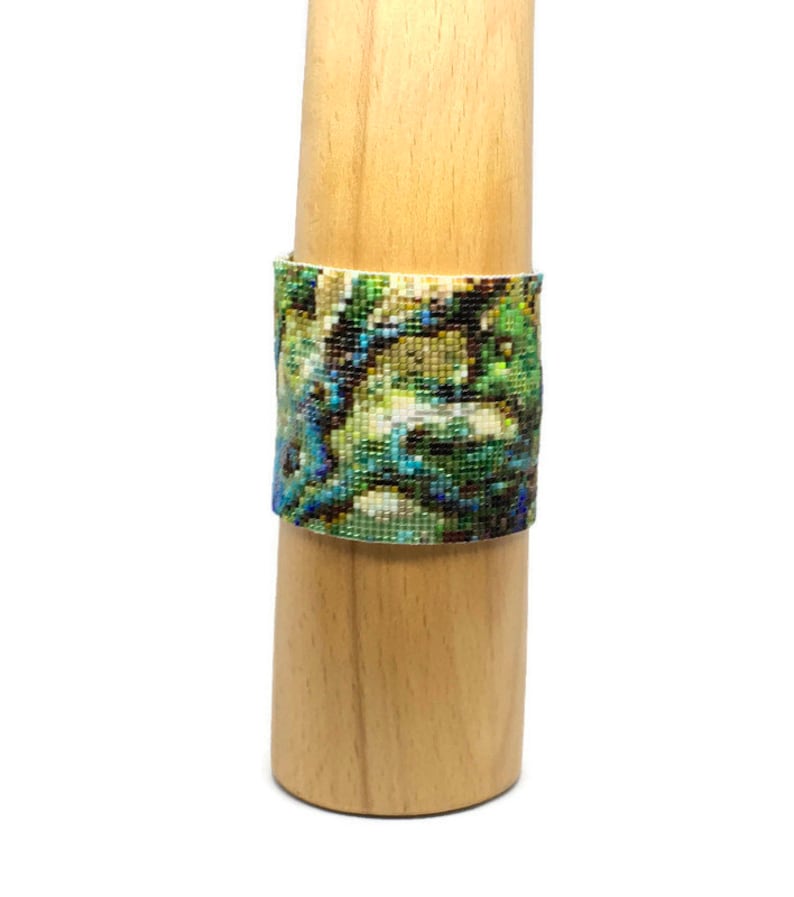 K&ouml;nnte beinhalten: Ein Armband aus Perlen mit einem farbenfrohen abstrakten Muster in Gr&uuml;n-, Blau- und Wei&szlig;t&ouml;nen. Das Armband wird auf einem Holzst&auml;nder pr&auml;sentiert.