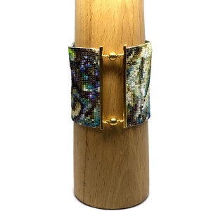 K&ouml;nnte beinhalten: Ein Armband aus Perlen mit einem farbenfrohen, abstrakten Muster. Das Armband hat einen goldenen Verschluss und wird auf einem Holzst&auml;nder pr&auml;sentiert.