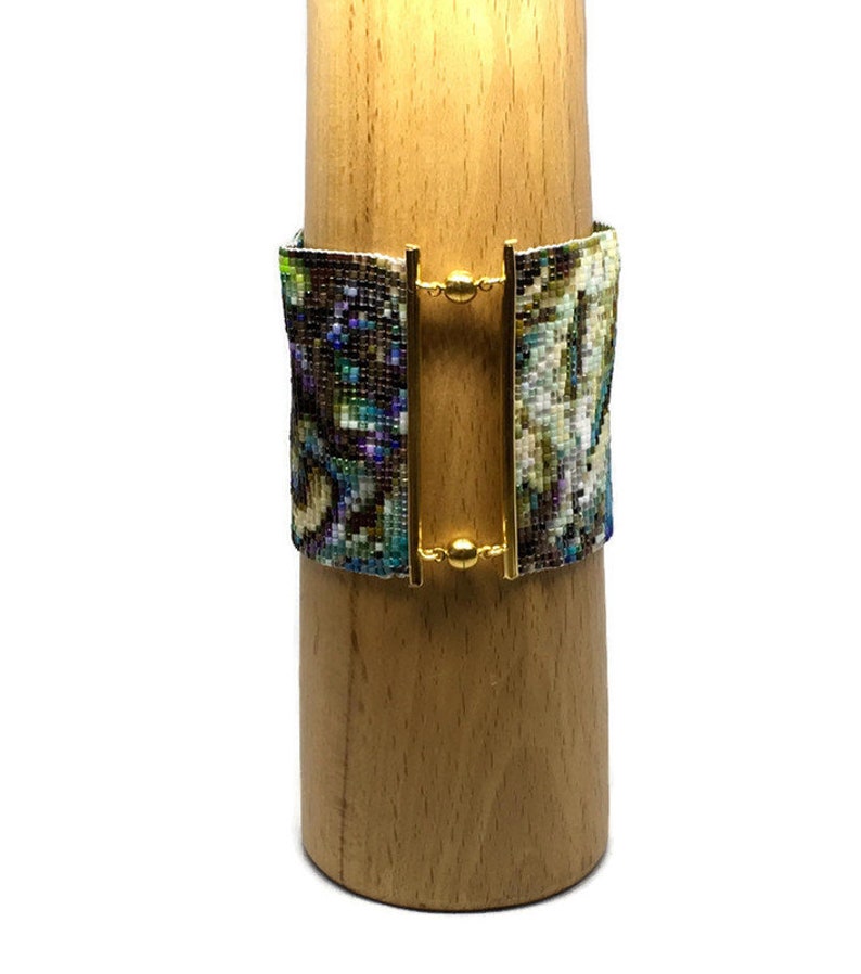 K&ouml;nnte beinhalten: Ein Armband aus Perlen mit einem farbenfrohen, abstrakten Muster. Das Armband hat einen goldenen Verschluss und wird auf einem Holzst&auml;nder pr&auml;sentiert.