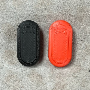 Pode incluir: Dois objetos ovais impressos em 3D. Um é preto, o outro é laranja brilhante. Ambos têm uma superfície texturizada e um design curvo na parte superior. Os objetos são provavelmente peças ou componentes pequenos.