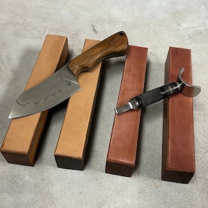 Op de afbeelding: Een handgemaakt mes met een houten handvat en een golvend lemmet ligt op een leren slijpblok. Een ander slijpgereedschap met een metalen lemmet en een zwarte handgreep is ook aanwezig, samen met extra slijpblokken.