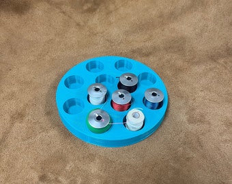 Può includere: Un organizer circolare turchese per rocchetti di filo che contiene diversi rocchetti di filo di vari colori. L'organizer ha più fessure circolari per contenere le bobine. Le bobine includono filo nero, rosso, bianco e blu.